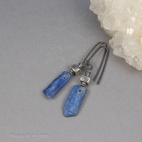 Blue stone earrings facing left showing raw kyanite's silvery sheen. Gray background.